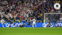 Deutscher Spieler schießt auf das Tor während eines Spiels gegen ein Team in blauen Trikots, vor einer vollen Tribüne.